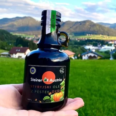 Styryjski olej z pestek dyni Steirer trzymany w dłoni na tle zielonej łąki i górskich krajobrazów Austrii.